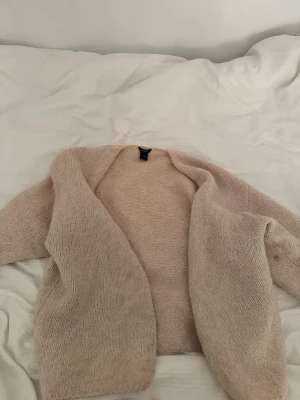 Beige stickad kofta från Lindex - Mysig beige stickad kofta från Lindex med öppen front och avslappnad passform. Koftan är gjord i ett mjukt material och har långa ärmar, perfekt för lager på lager-stil under kyliga dagar.