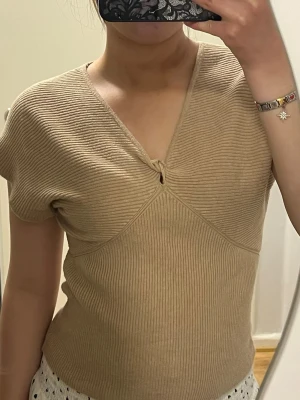 Beige ribbad v-ringad topp - Snygg beige ribbad topp med v-ringning och korta ärmar. Toppen har en twistdetalj framtill som ger en unik look och är tillverkad i ett mjukt, stretchigt material. Perfekt att styla med både jeans och kjol.