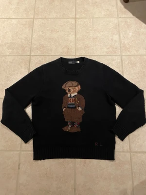 Svart stickad tröja med björn från Polo Ralph Lauren - Svart stickad tröja från Polo Ralph Lauren med ikonisk björn i brunt, beige och blått på bröstet. Tröjan har rund halsringning, ribbade muddar och RL-broderi nertill. Perfekt för dig som gillar klassiska och lekfulla detaljer.