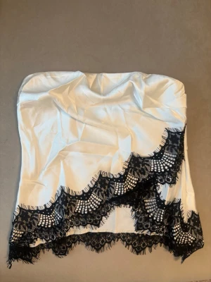 Vit axelbandslös topp med spets - Snygg axelbandslös topp i vit satin med svart spetsdetalj längs nederkanten. Toppen har en bandeauformad passning och är perfekt att styla med jeans eller kjol för en trendig look. Materialet är glansigt och känns lyxigt mot huden.