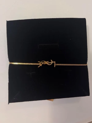 Guldfärgat armband från YSL - Snyggt och stilrent armband i guldfärg från Yves Saint Laurent med den ikoniska YSL-loggan som detalj. Kedjan är tunn och elegant, perfekt för att lyfta vilken outfit som helst. Kommer i en rosa ask.