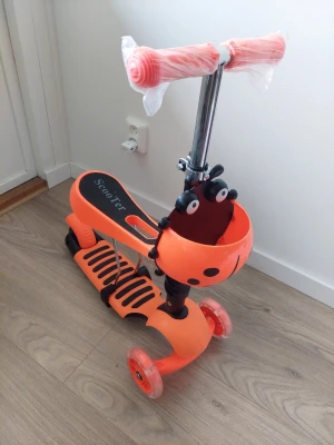 Orange sparkcykel med nyckelpigekorg - En orange och svart sparkcykel för barn med tre hjul och en söt korg framtill som ser ut som en nyckelpiga. Styret är justerbart i metall och har plastade handtag. Står 'Scooter' på fotplattan. Tillverkad i plast och metall.