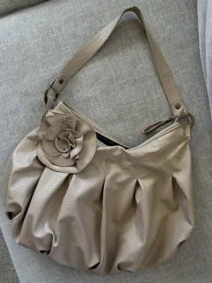 Beige axelväska med rosdetalj - Snygg beige axelväska i mjukt konstläder med en stor dekorativ ros framtill. Väskan har bred axelrem och dragkedja upptill. Perfekt för dig som gillar unika detaljer och vill ha en stilren men ändå personlig väska. Har en fläck på rosen. 