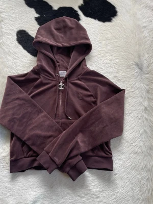 Brun velour hoodie från Juicy Couture i storlek L - Mysig brun hoodie i mjuk velour från Juicy Couture. Dragkedja framtill med ikonisk JC-berlock, huva med snören och känguruficka. Perfekt för en chill och trendig look. Broderad JC-logga på bröstet och ribbade muddar vid ärmslut och nederkant. Storlek L. 