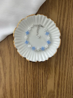 Sött armband med blå blommor och pärlor - Armband med små vita pärlor och blå blommor i plast. Silverfärgat justerbart spänne. Perfekt för dig som gillar gulliga och trendiga accessoarer med en lekfull touch.