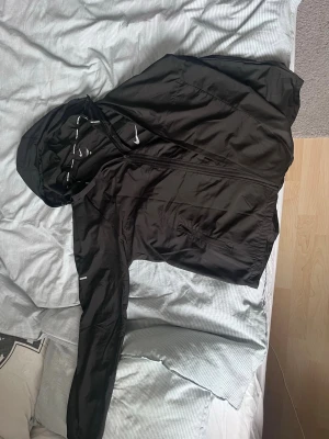 Svart Nike vindjacka med huva - Snygg svart vindjacka från Nike med huva och dragkedja framtill. Jackan har diskreta Nike-loggor och är tillverkad i lätt, vindtätt material. Perfekt för löpning eller när du vill ha en sportig look. Justerbar huva och praktiska fickor med dragkedja.