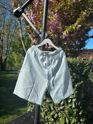 Randiga ljusblå shorts med snörning - Säljer ett par luftiga ljusblå shorts med vita ränder och snörning i midjan. Perfekta för varma dagar och har en avslappnad passform. Resår i midjan för extra komfort. Helt nya, för stora för mig.