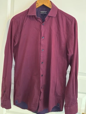 Mörkröd skjorta från Tommy Hilfiger - Snygg mörkröd skjorta från Tommy Hilfiger med klassisk krage och knappar framtill. Skjortan är långärmad och har en stilren design som passar perfekt till många olika outfits. Slim fit, 38/15.