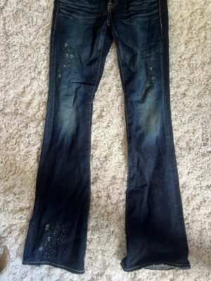 Mörkblå bootcut jeans med slitningar - Mörkblå jeans med bootcut-passform och tydliga slitningar samt ljusare partier framtill. Klassisk femficksmodell med kontrastsömmar och låg midja. Jeansen har en cool, använd look och är perfekta för en avslappnad stil.