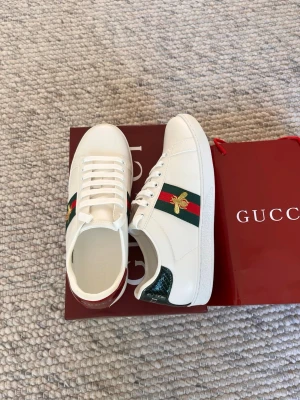 Gucci vita sneakers med bi-detalj - Säljer ett par vita sneakers från Gucci med klassiska gröna och röda ränder på sidorna samt en guldfärgad bi-detalj. Skorna har vita snören och detaljer i grönt och rött på hälen. Tillverkade i läder med gummisula. Perfekta för dig som gillar lyxiga och trendiga sneakers. Hör av dig innan du köper!