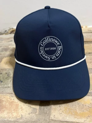 Mörkblå golf rope keps Golfinswe - Snygg mörkblå keps från Golfinswe med vit broderad logga framtill och texten 'Born in Sweden EST 2026'. Kepsen har en vit snodd längs skärmen och klassisk form. Perfekt accessoar för en sportig look.