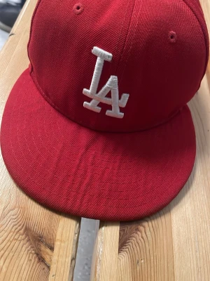 Röd LA-keps från New Era - Snygg röd keps från New Era med LA-logga broderad i vitt framtill. Klassisk flat brim och ventilerande hål upptill. Kepsen är gjord i bomullstyg och har en stilren streetwear-look. Perfekt för dig som gillar sportiga accessoarer.