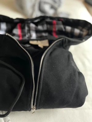 Svart hoodie från Burberry - Svart hoodie från Burberry med dragkedja och klassiskt rutigt foder i huvan. Broderad logga på bröstet och snörning i huvan. Perfekt för en stilren och avslappnad look.