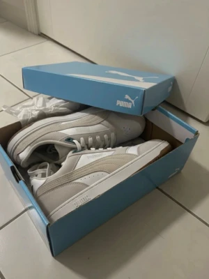 Vita och beige Puma sneakers - Snygga sneakers från Puma i vitt och beige med klassisk design. Ovandelen är i en mix av mocka och läder, och sulan är i gummi med ett greppvänligt mönster. Skorna har lågt skaft och vita snören. Perfekta för dig som gillar stilrena och tidlösa sneakers.