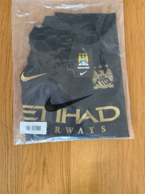 Svart Manchester City t-shirt Nike AGÜERO 16 - Svart t-shirt från Nike med Manchester Citys klubbmärke och stora guldfärgade tryck på bröstet. Snygg sportig stil med klassisk rund hals och korta ärmar. Perfekt för dig som älskar fotboll och vill visa ditt lagstöd. KÜN AGÜERO 16 på ryggen 