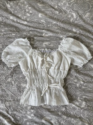 Vit blus med puffärm och knyt - Söt vit blus med puffärmar och broderade blommor. Toppen har smock i midjan och knyt framtill för en feminin look. Perfekt till jeans eller kjol för en trendig outfit.