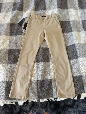 Beiga chinos från Replay - Barnstorlek 142cm/10A Snygga beiga chinos från Replay med klassisk rak passform och bälteshällor. Byxorna har två sidofickor och två bakfickor. Perfekta för en stilren och avslappnad look. Materialet är mjukt och bekvämt, troligtvis bomull med lite stretch.