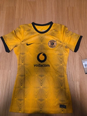 Nike Kaizer Chiefs 22/23 Dam, storlek XS - Gul fotbollströja med svart mönster och detaljer. Tröjan har klubbmärket Kaizer Chiefs och sponsorlogotypen Vodacom tryckta på framsidan. Nike-logotypen finns också på bröstet.