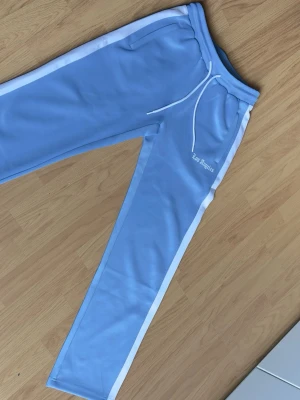 Black Squad babyblå tracksuit-byxor / wide leg strl S - Snygga babyblå tracksuit-byxor från Black Squad med vita stripes längs sidorna och “Los Angeles”-tryck framtill. Wide leg/passform med skönt material och justerbar midja. Storlek S. Perfekta för streetwear/Y2K-look. Bra skick.