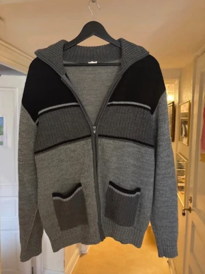 Stickad cardigan  - Snygg stickad ziptröja i grått och svart med huva och två framfickor. Tröjan har ribbade muddar och detaljer i olika grå nyanser samt svarta partier över axlarna. Perfekt för lager-på-lager och chill stil.