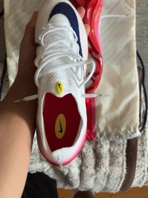 Nike Air Zoom Mercurial fotbollsskor - Snygga Nike Air Zoom Mercurial fotbollsskor i vitt med blå och rosa detaljer. Skorna har en modern design med snörning, röd sula och Nike-logga på både innersula och plös. Ovandelen är i mesh-liknande material för bra ventilation och lätt känsla.