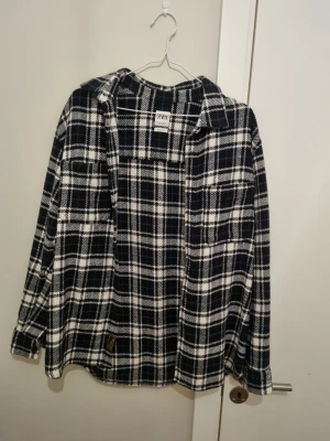Svartvit rutig skjorta från Zara - Snygg svartvit rutig skjorta från Zara med klassisk krage och långa ärmar. Skjortan har två bröstfickor och är tillverkad i ett mjukt tyg som känns skönt mot huden. Perfekt för en avslappnad och trendig look.