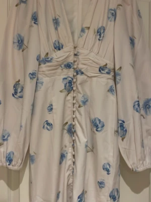 Vit långklänning med blå blommor - Romantisk långklänning i vitt med blått blommönster och v-ringning. Klänningen har långa puffärmar, knappar framtill och markerad midja. Perfekt för sommarens alla tillfällen. Klänningen är bara använd 2 gånger 