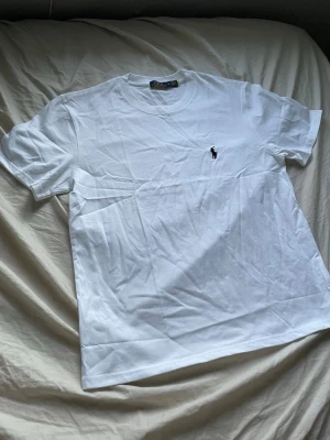 Vit t-shirt  - Klassisk vit t-shirt från Polo Ralph Lauren med rund halsringning och den ikoniska svarta logobroderingen på bröstet. Tillverkad i mjuk bomull och har en normal passform. Perfekt basplagg för en clean och stilren look.