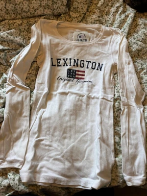 Vit långärmad topp från Lexington - Vit långärmad topp från Lexington med rund halsringning och tryck framtill med Lexington-logga och flagga. Tillverkad i mjuk bomull, perfekt för en avslappnad och stilren look.