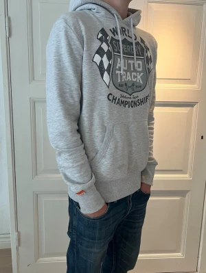 Grå Superdry hoodie med tryck - Snygg ljusgrå hoodie från Superdry med stort svart och vitt tryck på bröstet med racingtema. Klassisk känguruficka framtill, dragsko i huvan och ribbade muddar. Perfekt för dig som gillar streetwear och sportig stil.