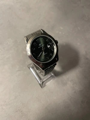 Silverfärgad Riccoco klocka med arabisk urtavla - Stilren klocka från Riccoco med silverfärgat armband i metall och mörkgrön urtavla. Urtavlan har arabiska siffror och datumvisning. Klassisk rund form och länkarmband som ger en cool vibe. Perfekt för dig som gillar detaljer och vill sticka ut med din accessoar. Saknar övre del på armbandet. 