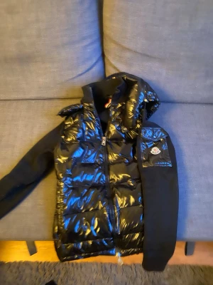 Svart dunväst med huva från Moncler - Snygg svart dunväst från Moncler med glansig yta och huva. Västen har dragkedja framtill, stickade svarta ärmar och Moncler-logga på vänster ärm. Perfekt för lager-på-lager och streetstyle.