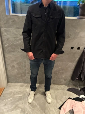 Svart skjorta med knappar - Stilren svart overshirt skjorta knappar framtill. Skjortan har två bröstfickor med lock och klassisk krage. Passar perfekt till jeans för en avslappnad look.