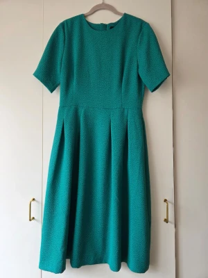 Grön klänning med struktur HM - En stilren grön klänning med rund halsringning och korta ärmar. Klänningen har en markerad midja och veckad kjol som ger en fin siluett. Materialet är strukturerat. Klassisk look.