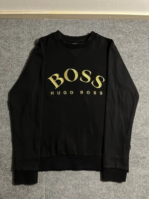 BOSS Hugo Boss – Black Sweatshirt (M) - Rare and stylish BOSS Hugo Boss sweatshirt in classic black. Size M, length 63 cm, width 50 cm – fits perfectly for a clean, modern silhouette. The front features the iconic BOSS HUGO BOSS logo in elegant gold print, giving it a premium and timeless look.  ✅ Condition: Excellent – no wear, no stains 💯 Authenticity: 100% genuine, original Hugo Boss branding 📦 Shipping: Sent immediately after purchase – fast and reliable delivery!  This sweatshirt is hard to find nowadays