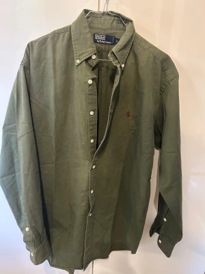 Olivgrön skjorta från Polo Ralph Lauren - Klassisk olivgrön skjorta från Polo Ralph Lauren med knappslå och vit knappdetalj. Skjortan har lång ärm, button-down krage och den ikoniska lilla röda loggan broderad på bröstet. Perfekt till jeans eller chinos för en clean look. Den är lite större i storleken så den passar bäst om man vanligtvis har M i storlek