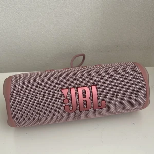 JBL Flip 6 rosa bärbar högtalare - Snygg rosa JBL Flip 6 bärbar högtalare med robust tygmaterial och cylinderformad design. Har tydliga knappar för volym, power och Bluetooth samt USB-C laddningsport. Perfekt för att ta med sig musiken överallt.