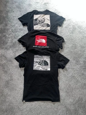 Svarta The North Face t-shirts - Tre svarta t-shirts från The North Face med tryck på ryggen och liten logga på bröstet. Två har grafiskt svartvitt tryck och en har röd logga med texten 'Never Stop Exploring'. Klassisk passform och rund hals. Perfekta för en stilren och avslappnad look.