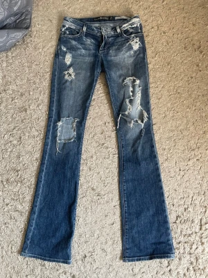 Blå slitna bootcut-jeans från Dylan George  - ett par skit snygga blå bootcut-jeans från Dylan George med slitna detaljer och hål på benen. Jeansen har klassisk femficksdesign, låg midja och är gjorda i denim. Perfekta för dig som gillar en edgy och avslappnad stil. Dem hade ingen framknapp när jag köpte så har fixat de själv.  