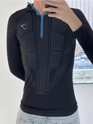 Gymshark V1 - Säljer en svart långärmad träningströja från Gymshark med blå detaljer och half zip. Tröjan har ett sportigt mönster framtill och huva med blå kant. Perfekt för gymmet