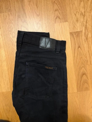 Svarta jeans från Nudie Jeans - Säljer ett par svarta jeans från Nudie Jeans med klassisk femficksdesign och diskret logga på bakfickan. Jeansen har en smal passform och är tillverkade i mjukt denimtyg som sitter snyggt. Perfekta för en clean och stilren look.