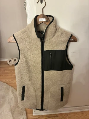 Beige fleeceväst med svarta detaljer - Mysig beige fleeceväst med svarta detaljer och dragkedja framtill. Västen har en hög krage, två fickor med svarta kanter och en svart bröstficka med dragkedja. Perfekt för lager-på-lager och chill stil.