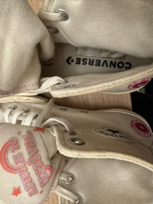 Converse Chuck Taylor vita high tops - Säljer ett par vita Converse Chuck Taylor All Star high tops med klassisk svart stjärnlogga på sidan och röda detaljer på tungan. Skorna är tillverkade i canvas och har metallöljetter för snörningen. Perfekt för dig som gillar ikoniska sneakers.
