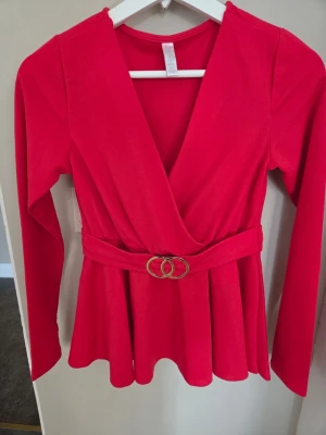 Röd omlottopp med bälte - Snygg röd omlottopp med lång ärm och markerad midja tack vare ett matchande bälte med guldfärgat spänne. STL XS Toppen har v-ringad omlottskärning och ett lätt utställd nederdel för en feminin siluett. Perfekt för dig som vill sticka ut med färg. FRÅN NEW YORKER I MÄRKET AMISU , ANV 1 G 