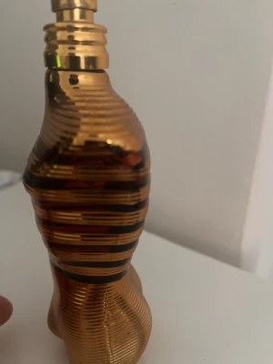 Jean Paul Gaultier Le Male parfym - Unik parfymflaska från Jean Paul Gaultier formad som en manlig torso i metallic guld med svarta ränder. Flaskan har en modern och lyxig design som verkligen sticker ut på hyllan. Perfekt för dig som gillar exklusiva och ikoniska dofter. Den innehåller 100ml kvar i flaskan! 