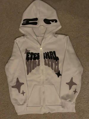 Vit hoodie med tryck och huva - Säljer en vit hoodie med dragkedja och stor huva. På bröstet står det 'ETERNAL WORTH' i svart och grått tryck. Ärmarna har stjärnliknande motiv i grått och svart. Huvan har svarta grafiska detaljer. Perfekt för dig som gillar streetwear och unik design.  Ordinarie pris var 799 kr.Passar dig som är runt 150 cm lång men hör av er om frågor kring storlek och annat! 😁