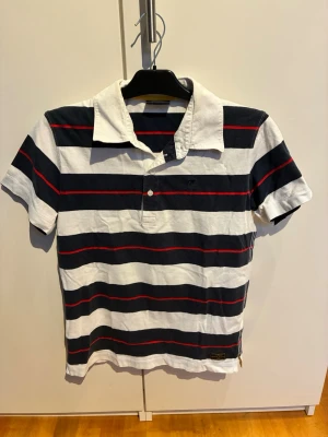 Peak performance pikétröja  - Snygg pikétröja med breda marinblå och vita ränder samt tunna röda detaljer. Klassisk krage och kort ärm, perfekt för en avslappnad stil. Tillverkad i mjuk bomull och har knappslå framtill.