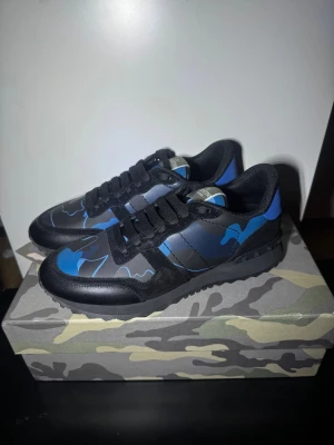 Rock runners - Snygga sneakers från Valentino i svart och blått med camouflagemönster. Skorna har lågt skaft, svarta snören och en robust sula med mönstrad undersida. Ovandelen är i läder och textil, vilket ger en cool och modern look.