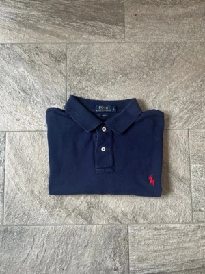 Marinblå pikétröja från Polo Ralph Lauren - Klassisk marinblå pikétröja från Polo Ralph Lauren med röd broderad logga på bröstet. Tröjan har krage, knappar framtill och är tillverkad i bomull för en skön och avslappnad stil.