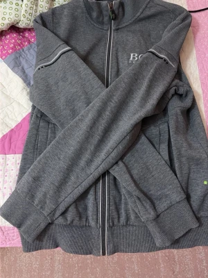 Grå zip hoodie från BOSS - Snygg grå hoodie från BOSS med dragkedja framtill och diskret logga på bröstet. Tröjan har ribbade muddar, ståkrage och två fickor på sidorna. Tillverkad i mjukt material som soppar in en sportig vibe. Perfekt för dig som gillar stilrena och bekväma plagg.